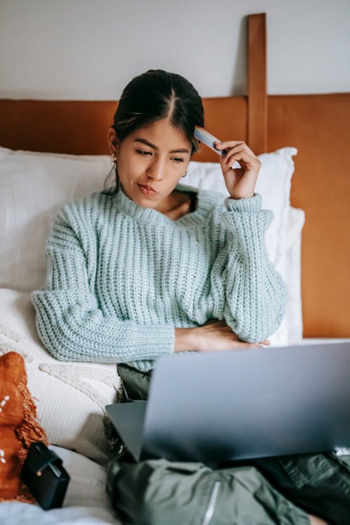 Young ethnic focused female confirming information of credit card online in netbook while resting on bed